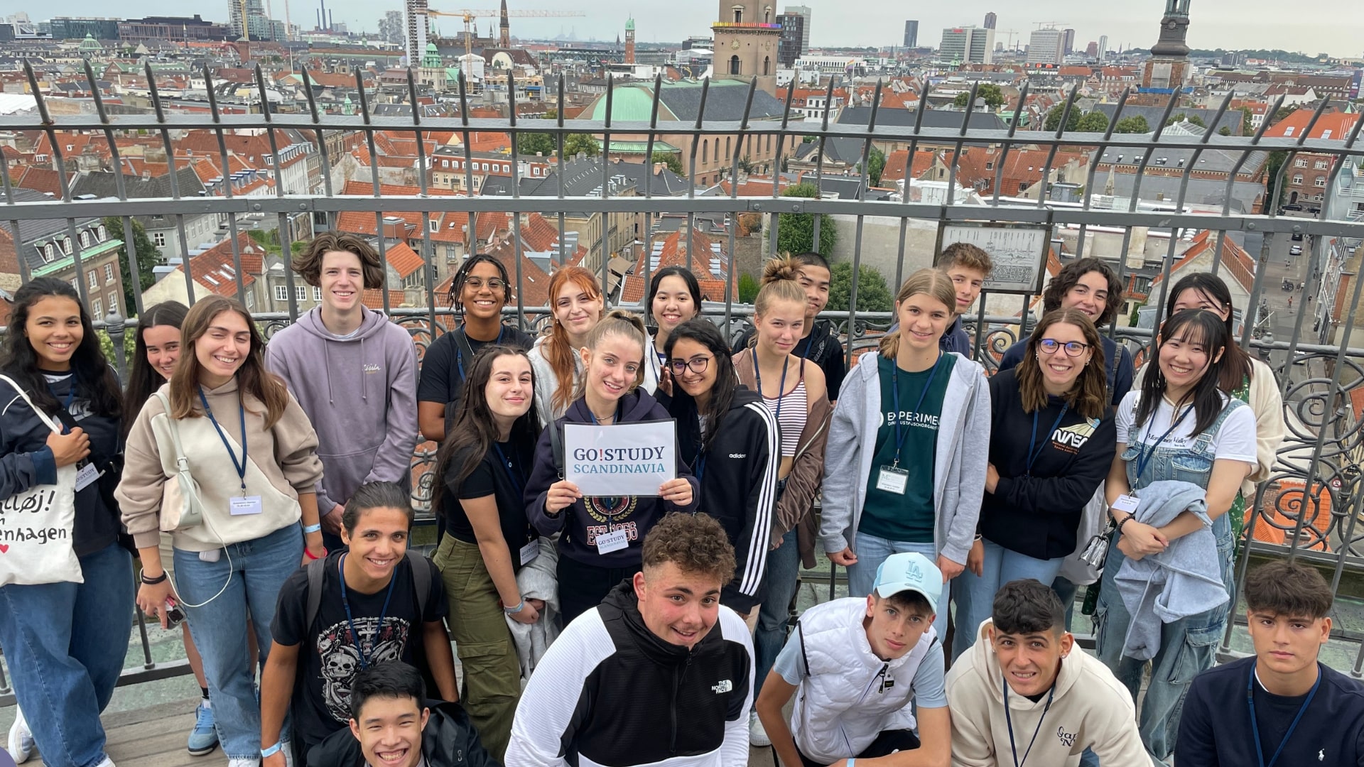 Un gruppo di studenti sorridenti in posa su una terrazza panoramica, con una vista della città di Copenaghen sullo sfondo. Al centro, una studentessa tiene un cartello con la scritta "Go! Study Scandinavia