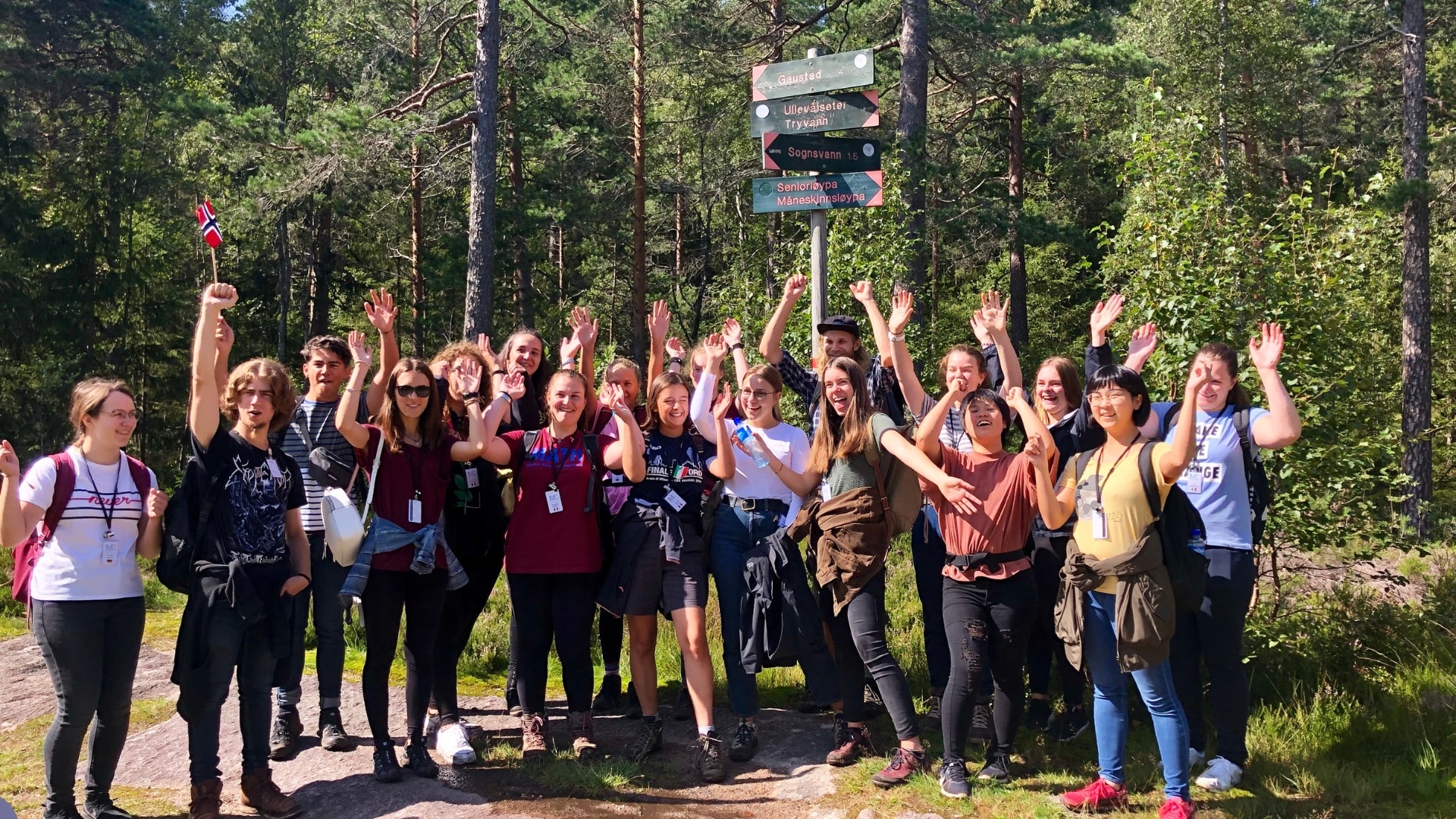 Un gruppo di giovani sorridenti posa all'aperto in una foresta, alzando le mani e salutando verso la fotocamera. Alcuni indossano zaini, e una persona tiene una piccola bandiera norvegese. 