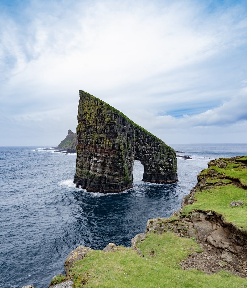 Una formazione rocciosa spettacolare a forma di arco nell'oceano vicino alla costa verdeggiante delle Isole Faroe, con un cielo parzialmente nuvoloso.