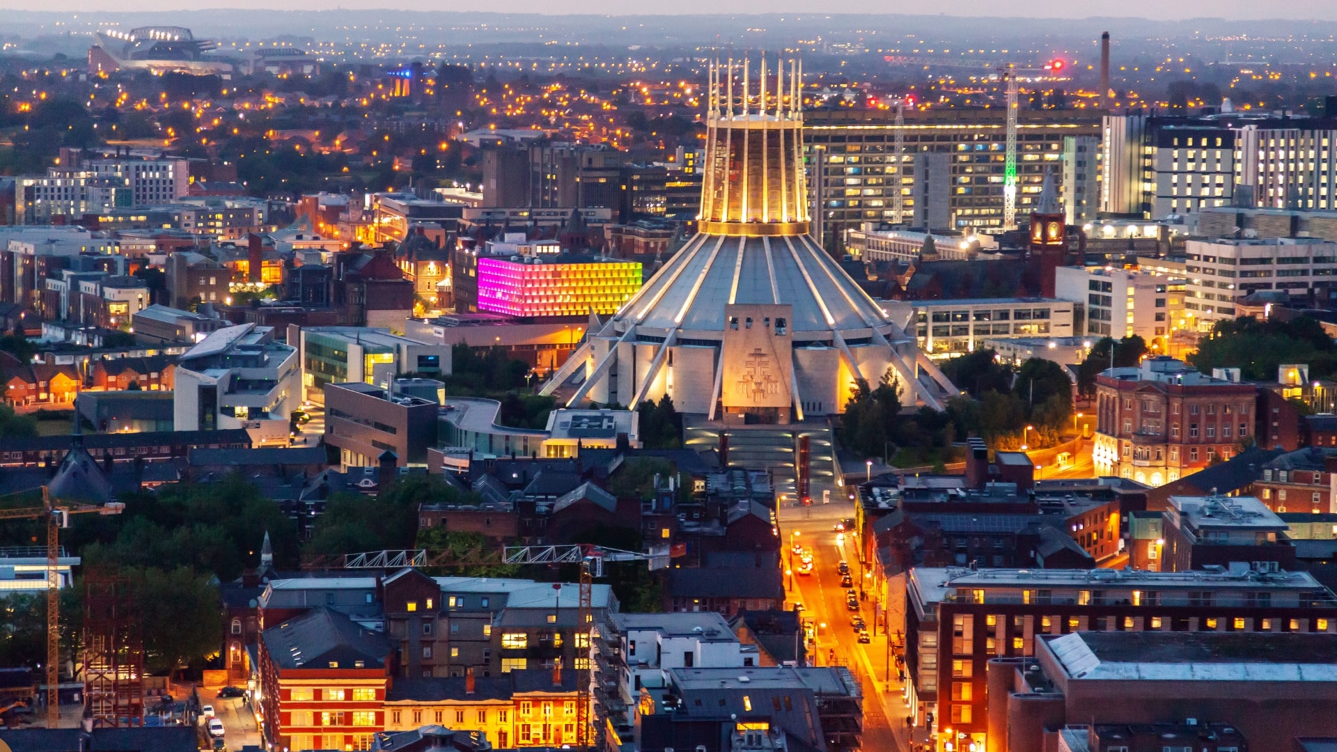 Vista notturna di Liverpool con luci accese, dominata al centro dalla Cattedrale Metropolitana di Cristo Re, un edificio moderno a forma circolare con guglie appuntite