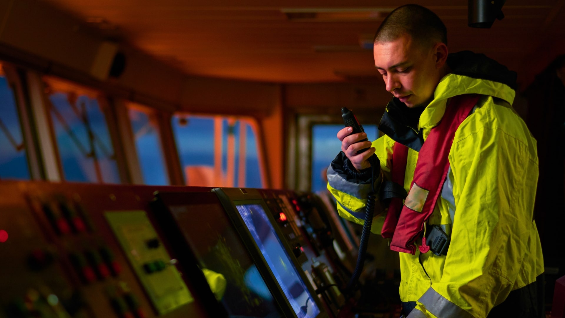 Marinaio nella cabina di comando di una nave che comunica tramite radio, con giacca impermeabile gialla e giubbotto di salvataggio rosso, circondato da strumenti di navigazione.